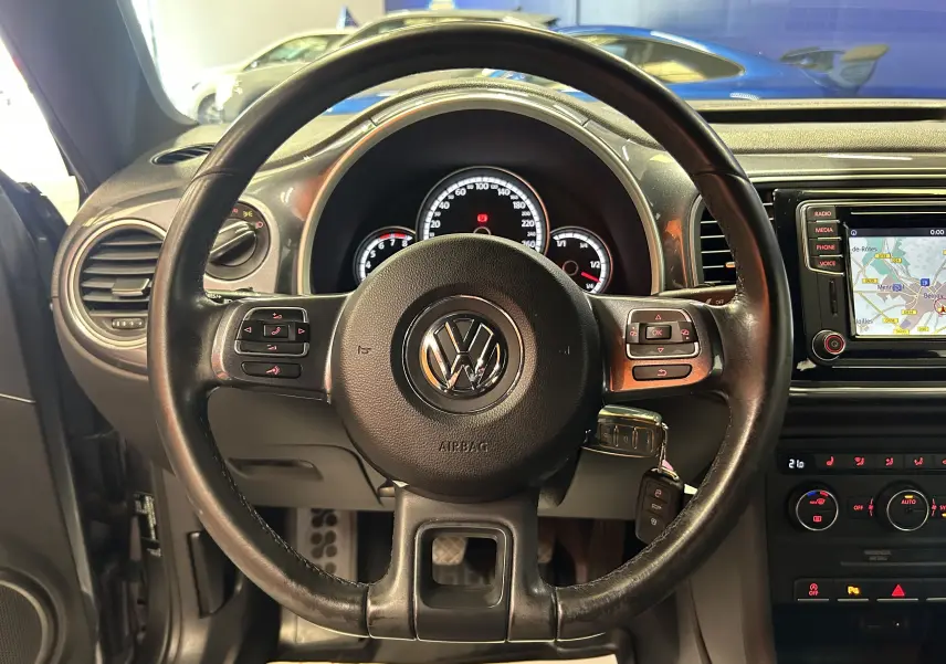 Vue intérieure centrée sur le volant en cuir noir d'une Volkswagen Coccinelle 2016 avec écran GPS allumé.