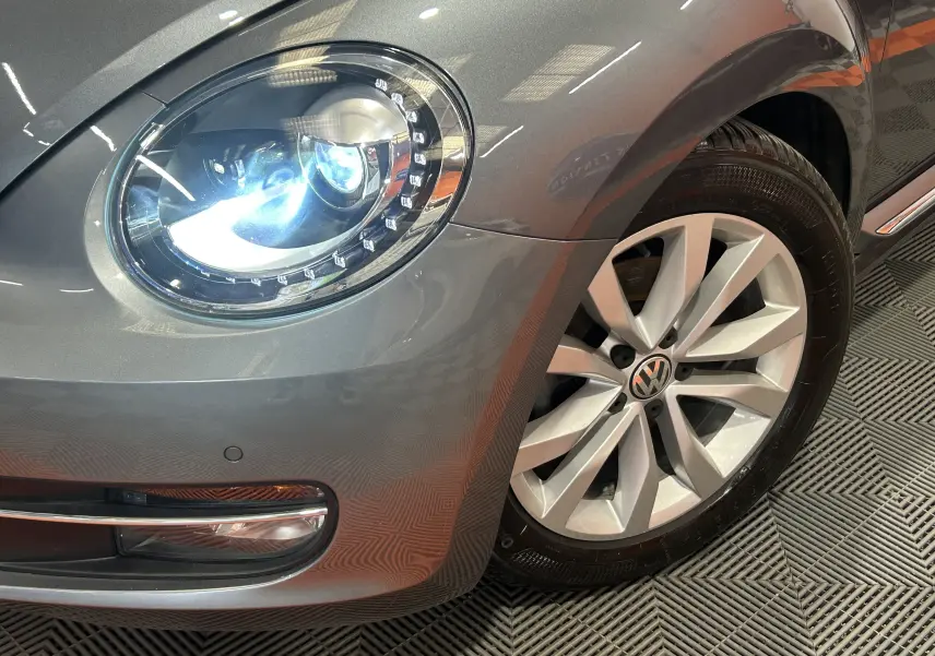 Gros plan sur l'avant droit gris métallisé d'une Volkswagen Coccinelle 2016, mettant en valeur le phare LED et la jante alliage.