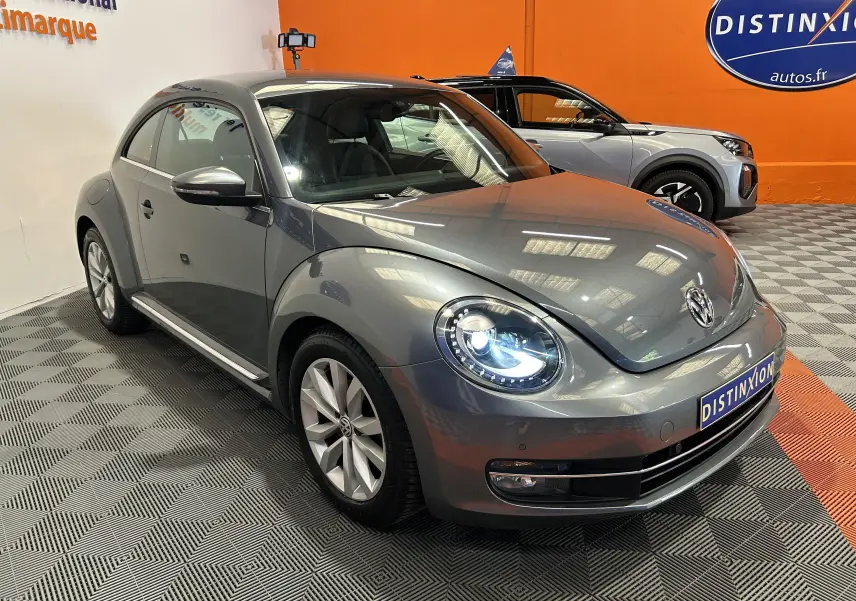 Volkswagen Coccinelle gris vue 3/4 avant droit avec phares allumés dans un showroom au sol à damier.