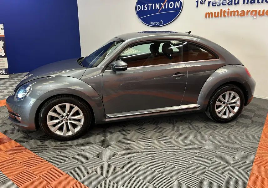Vue de profil côté gauche d'une Volkswagen Coccinelle gris métallisé, avec jantes alliage et phares allumés, en intérieur showroom.