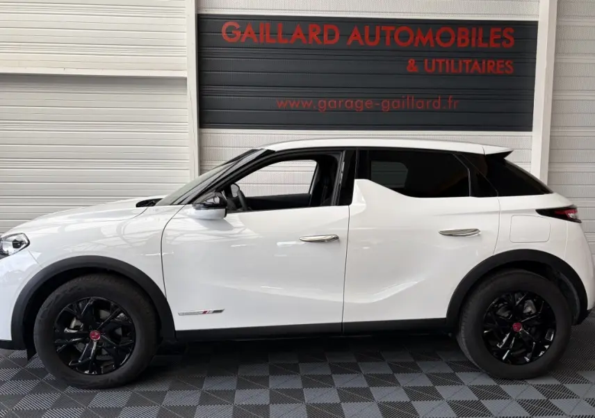 Vue latérale droite d'un DS3 Crossback blanc avec jantes noires et vitres arrière teintées, en intérieur showroom.