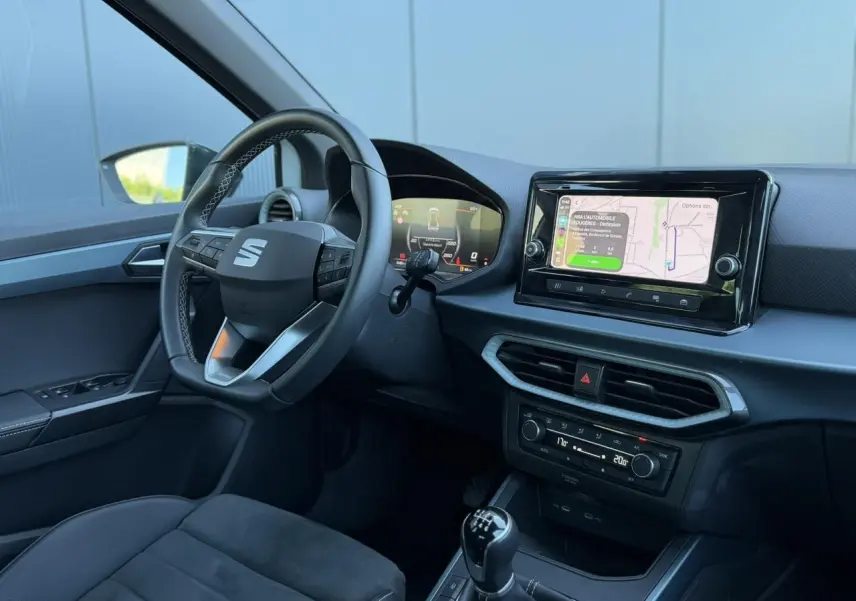 Vue intérieure côté conducteur du SEAT Arona TSI 115 Xpérience 2025, avec volant multifonction, écran tactile et levier de vitesse manuel.
