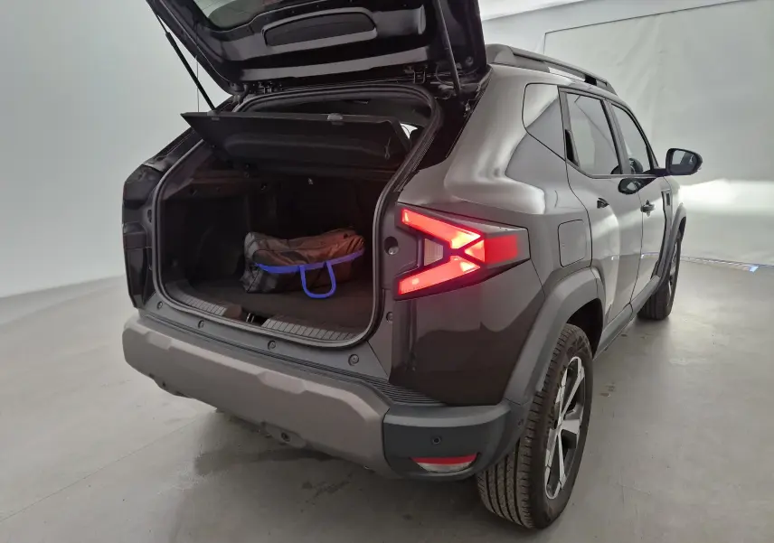 Vue 3/4 arrière droite du Dacia Duster noir 2024 avec coffre ouvert et sac à l'intérieur, feux arrière allumés.