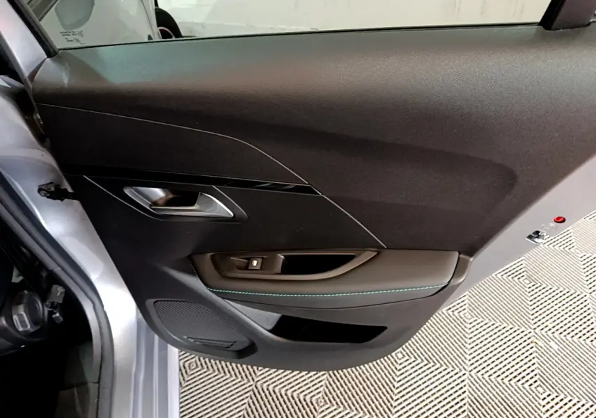 Vue rapprochée de la porte avant gauche gris clair de la Peugeot 208 PureTech 100 S&S Allure Business avec poignée argentée et surpiqûres bleues.
