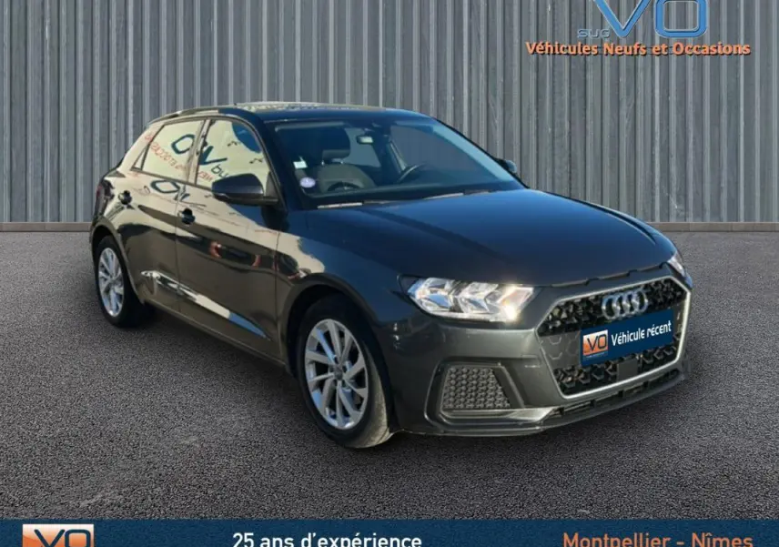 Audi A1 Sportback gris foncé vue 3/4 avant droit avec phares allumés sur fond urbain neutre.