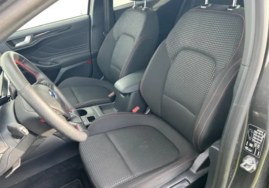 Vue intérieure côté conducteur de la Ford Focus IV ST-Line X, sièges tissu noir avec surpiqûres rouges et volant sport.