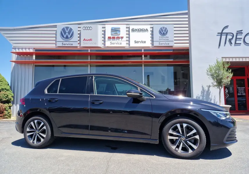 Profil côté gauche d'une Volkswagen Golf VIII noire métallisée stationnée devant un garage multimarque.