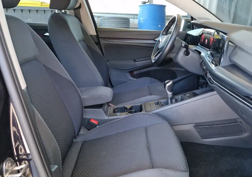 Intérieur côté conducteur d'une Volkswagen Golf VIII noire, sièges tissu gris et tableau de bord moderne avec boîte manuelle.