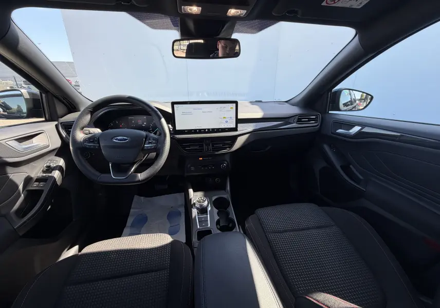 Vue intérieure avant de la Ford Focus IV ST-Line noire, tableau de bord moderne avec écran tactile central et volant sport à surpiqûres rouges.