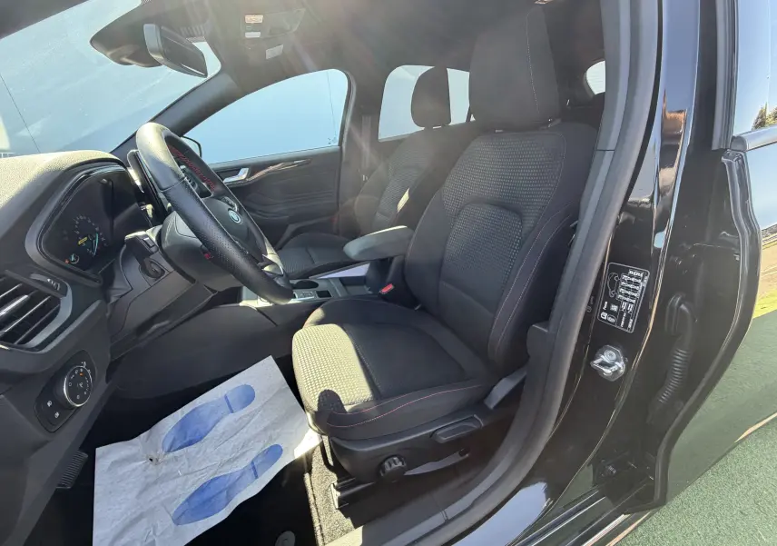 Vue intérieure côté conducteur de la Ford Focus IV ST-Line noire avec sièges tissu et surpiqûres rouges visibles.