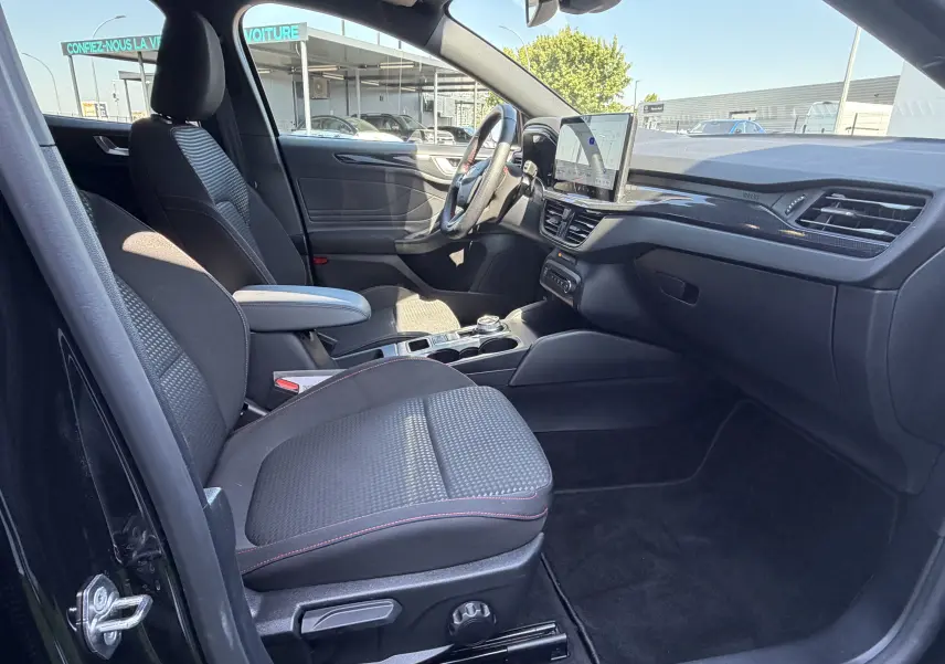 Vue intérieure côté conducteur de la Ford Focus IV ST-Line noire, avec sièges tissu et surpiqûres rouges, tableau de bord moderne.