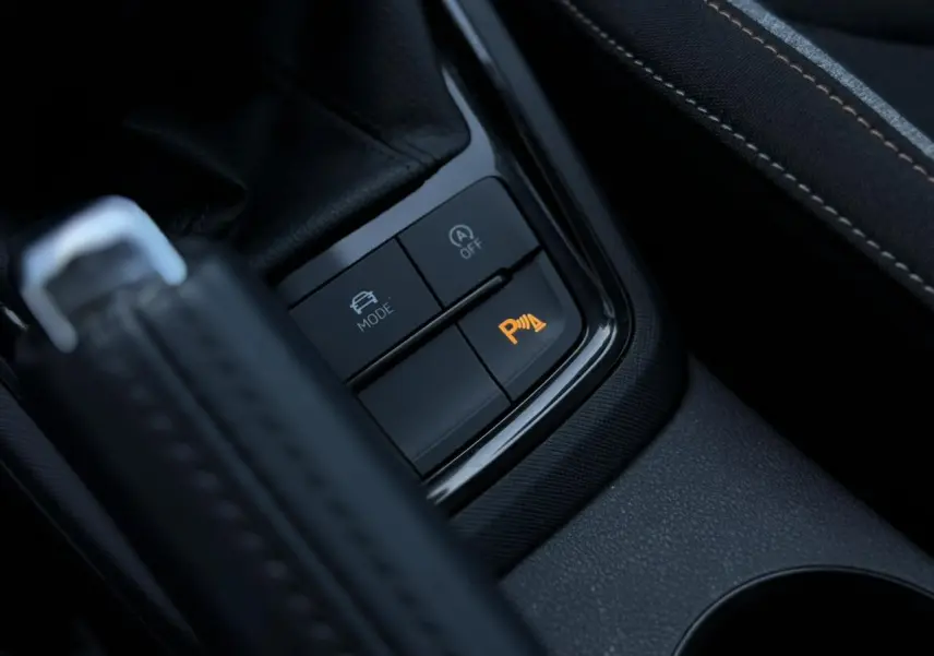 Gros plan sur les commandes de la console centrale avec frein à main et bouton orange d’aide au stationnement actif.
