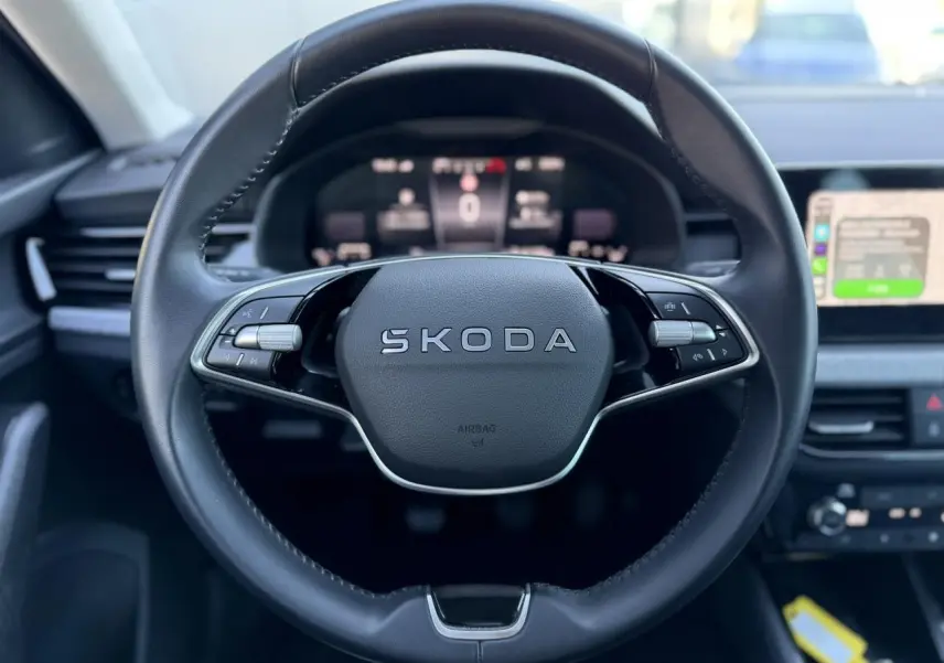 Gros plan sur le volant en cuir noir avec logo SKODA, tableau de bord numérique et écran tactile à droite.