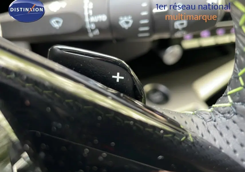 Gros plan sur la palette de changement de vitesse "+" et partie du volant cuir noir avec surpiqûres vertes du Peugeot 308 gris Artense.