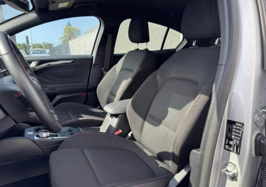 Vue intérieure côté gauche de la Ford Focus IV ST-Line X 2025, sièges tissu gris avec surpiqûres rouges visibles.