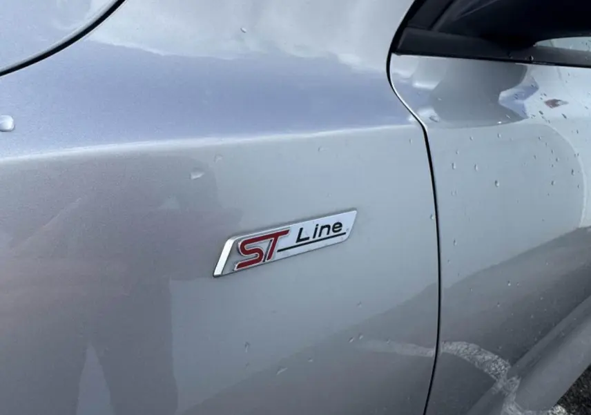 Gros plan sur le badge latéral ST-Line de la Ford Focus IV gris Moondust Metal, côté gauche avec gouttes d'eau.