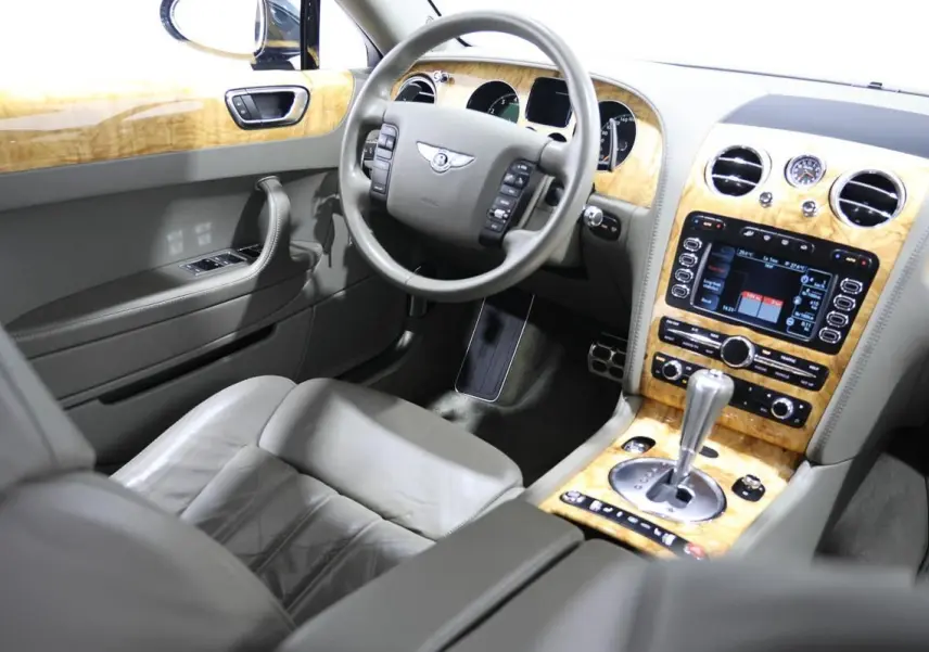 Vue intérieure côté conducteur de la Bentley Continental Flying Spur 2006 avec tableau de bord en bois clair et cuir gris.