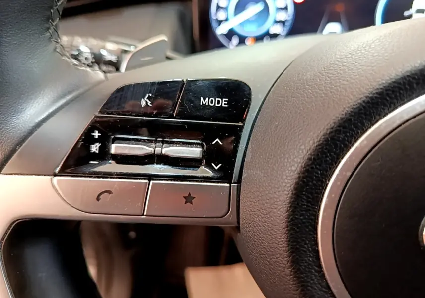 Gros plan sur les commandes gauche du volant cuir noir du Hyundai Tucson 1.6 Hybrid 230, tableau de bord numérique en arrière-plan.