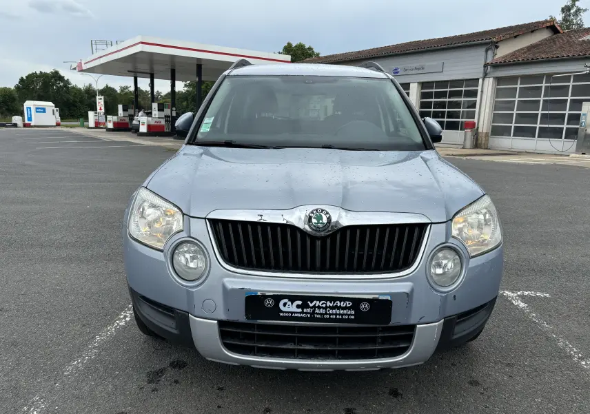 Vue frontale d'un Skoda Yeti gris 2012 avec calandre noire et phares allumés sur parking devant station-service.