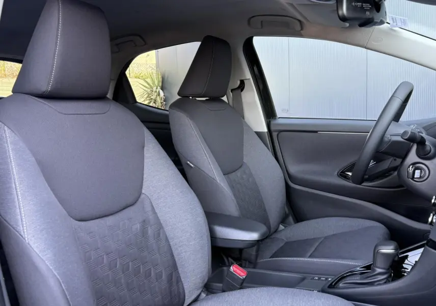 Vue intérieure côté conducteur de la Toyota Yaris 2025 hybride, sièges gris avec accoudoir central et volant cuir.