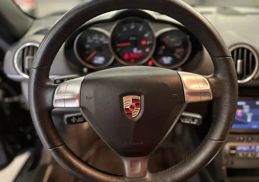 Intérieur de Porsche Cayman 987 noir, vue rapprochée du volant cuir avec commandes Tiptronic et tableau de bord en arrière-plan.