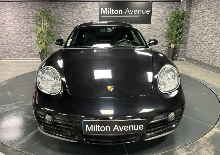 Vue avant d'une Porsche Cayman noire 2008 avec phares ronds et plaque Milton Avenue en intérieur showroom.