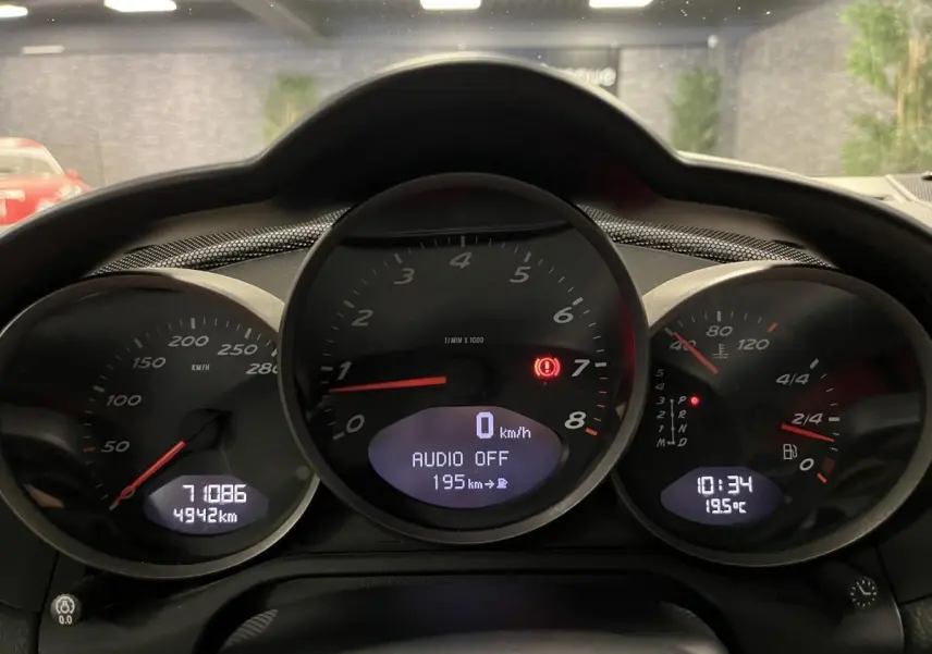 Gros plan sur le tableau de bord noir du Porsche Cayman 987 avec compte-tours central et affichages numériques.