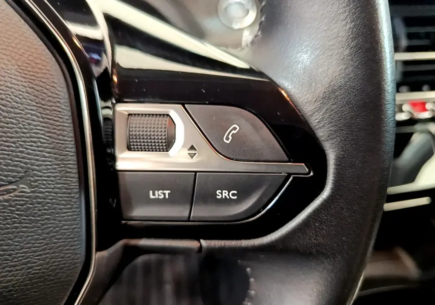 Gros plan sur les commandes du volant côté gauche en cuir noir du Peugeot 208 blanc PureTech 100 S&S Allure Business.