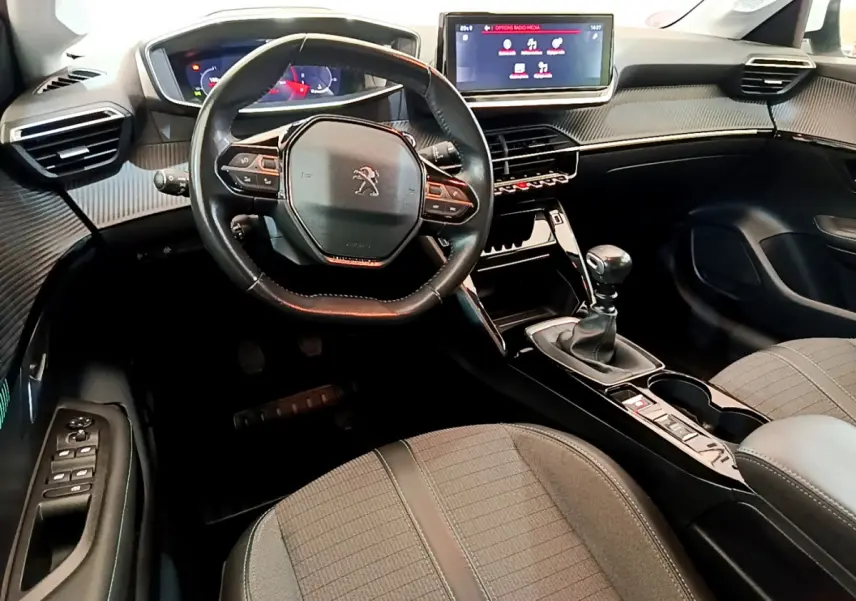 Vue intérieure avant du Peugeot 208 blanc, volant cuir compact, écran tactile 10 pouces et boîte manuelle visible.