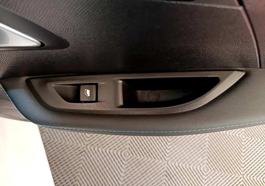 Gros plan sur la commande de lève-vitre côté gauche intérieur d'une Peugeot 208 blanche avec surpiqûres bleues.