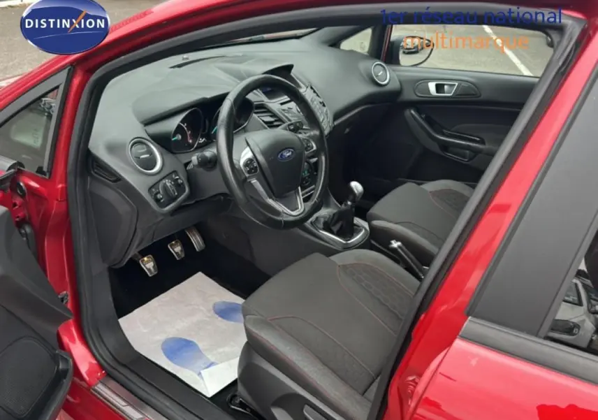 Vue intérieure côté conducteur d'une Ford Fiesta rouge 2017 avec volant cuir, boîte manuelle et sièges tissu noirs.