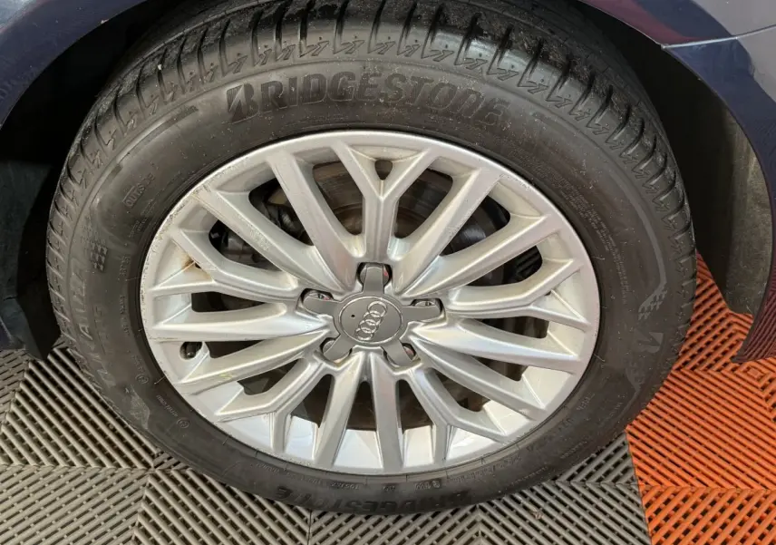 Gros plan sur la jante alliage argentée avec logo Audi d'une Audi A3 bleu, pneu Bridgestone visible au-dessus d'un sol carrelé.