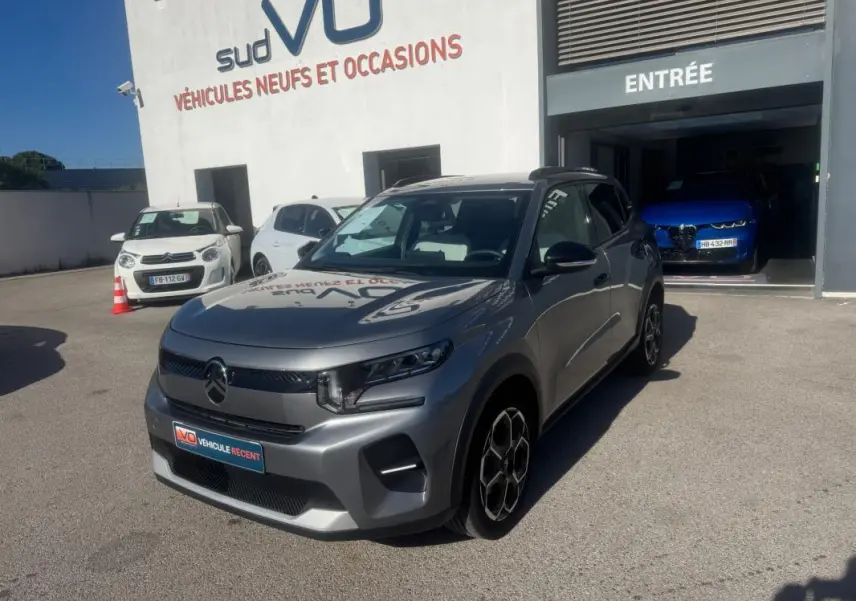 Citroën C3 gris 3/4 avant droit avec jantes alliage et barres de toit, stationnée devant un garage.