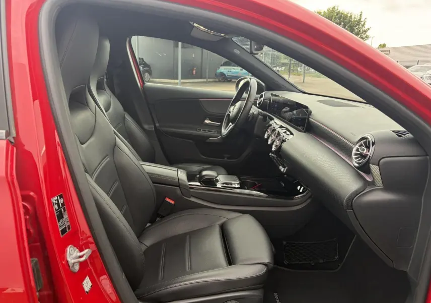 Vue intérieure côté conducteur d'une Mercedes Classe A rouge, avec sellerie cuir noir et tableau de bord moderne.