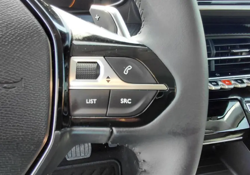 Gros plan sur les commandes gauche du volant cuir noir de la Peugeot 208 Hybrid 110 ch, avec tableau de bord et aérateurs en arrière-plan.