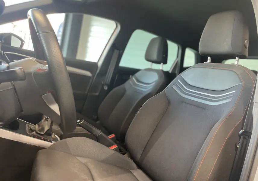 Vue intérieure côté conducteur du SEAT Arona 2024, sièges textile noirs avec surpiqûres orange et volant sport cuir.