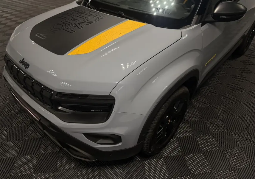 Vue 3/4 avant droit d’un Jeep Avenger gris avec stickers The North Face noirs et jaunes sur le capot et jantes noires.