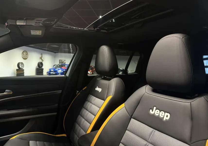 Vue intérieure des sièges avant noirs et gris avec surpiqûres jaunes et logo Jeep, toit ouvrant en verre visible au-dessus.