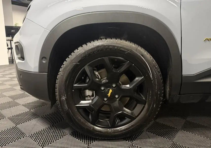 Gros plan sur la roue avant gauche noire avec jante alliage noir mat et partie de la carrosserie gris clair du Jeep Avenger.