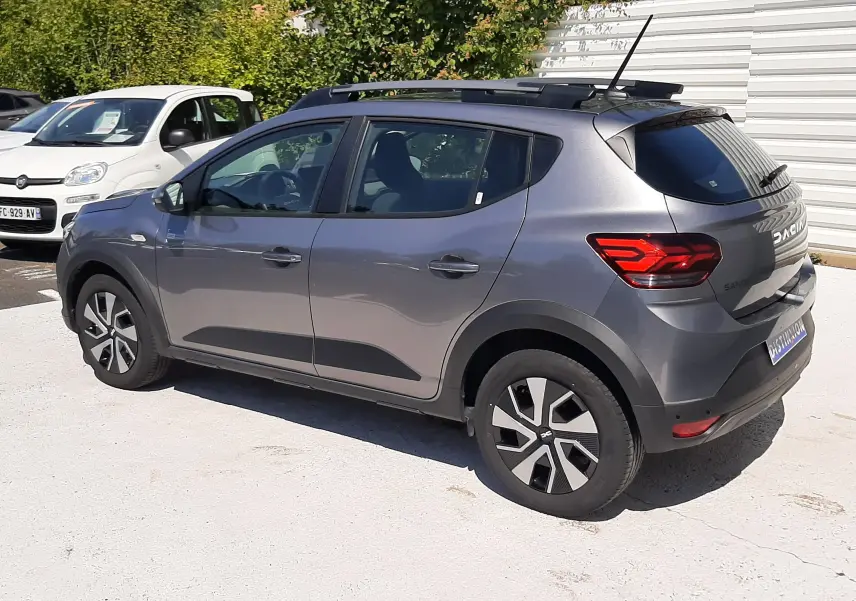 Dacia Sandero Stepway gris schiste vue 3/4 arrière droit, avec barres de toit et jantes spécifiques.
