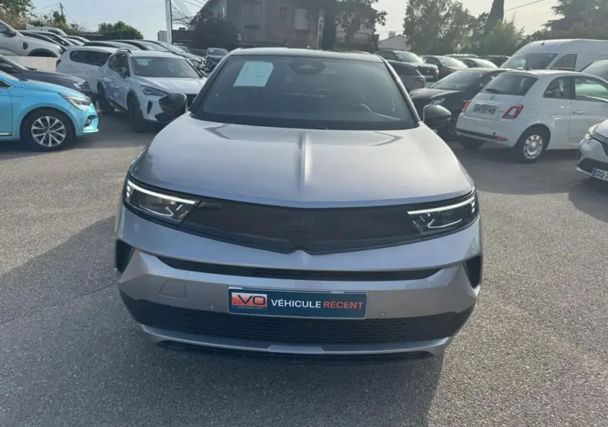 Vue frontale d'un Opel Mokka électrique gris 2025 avec calandre noire et phares LED fins, sur parking extérieur.