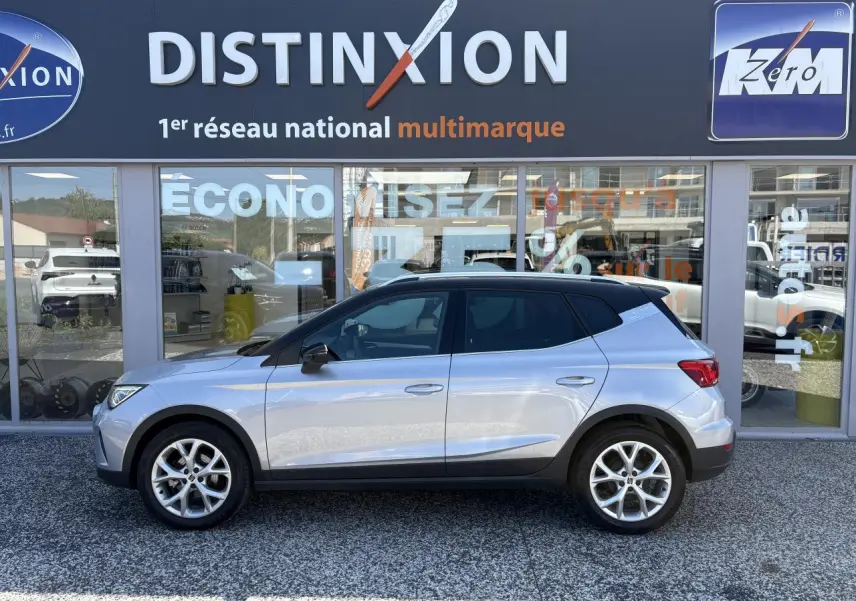 Vue de profil côté gauche d'un SEAT Arona 2024 gris clair avec jantes alliage et toit noir devant un showroom multimarque.