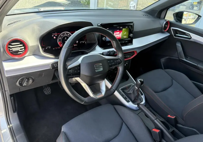 Vue intérieure côté conducteur du SEAT Arona 2024, volant sport cuir noir, inserts rouges autour des aérateurs et console centrale.