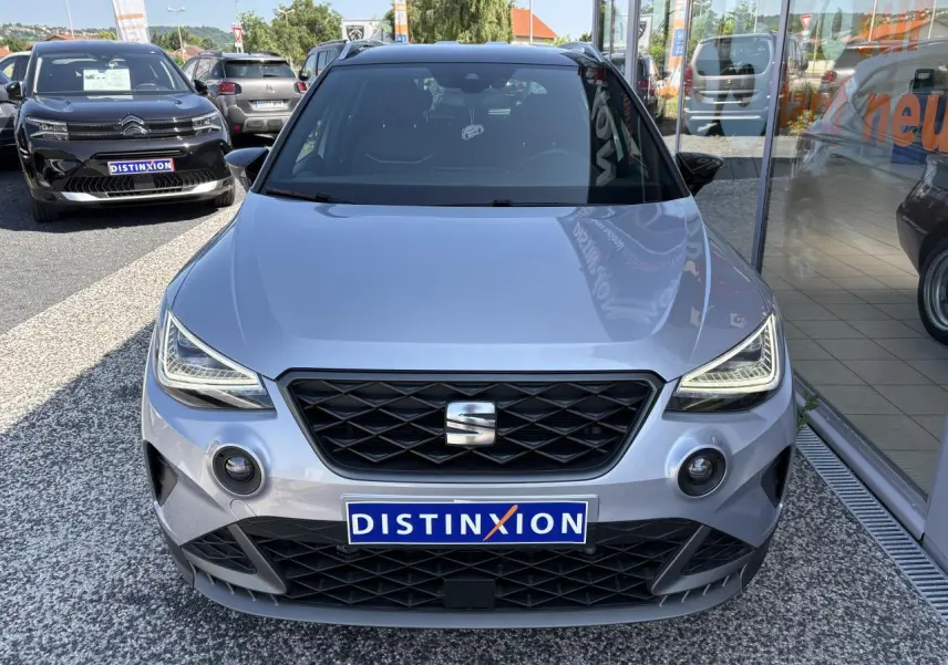 Vue avant d'une SEAT Arona 2024 gris clair, avec calandre noire et phares LED anguleux, sur parking extérieur.