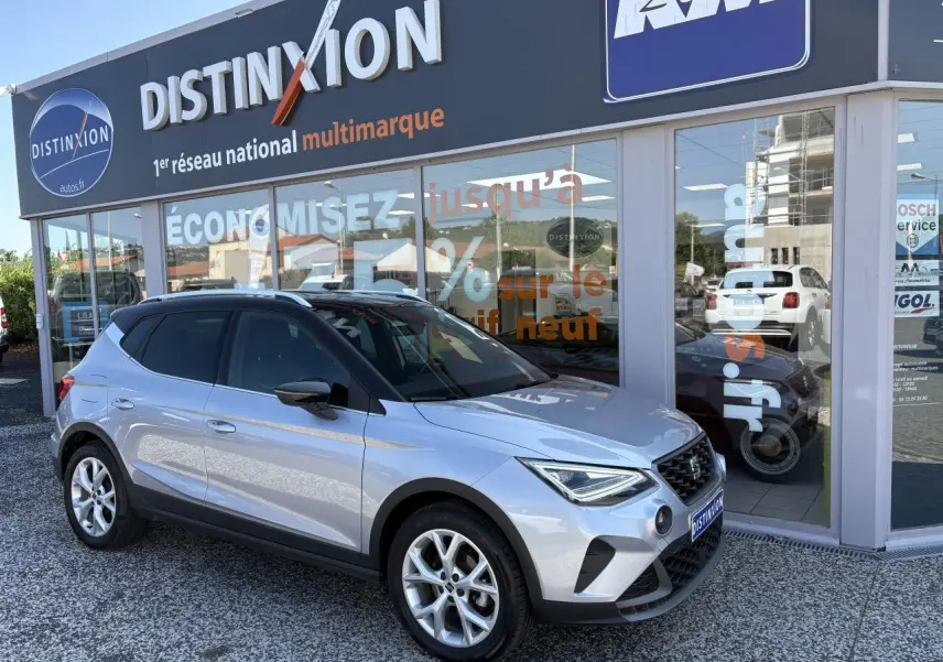 SEAT Arona 2024 gris clair en 3/4 avant droit, avec jantes alliage et toit noir devant concession multimarque.