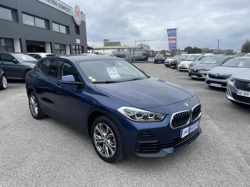 BMW X2 X-Drive 20 da 2021 bleu métal vue 3/4 avant droit sur parking de voitures en concession.