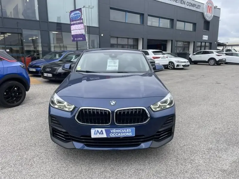 Vue avant d’un BMW X2 bleu métal 2021 avec calandre double haricot et phares allumés sur parking de concession.