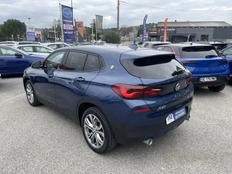 BMW X2 bleu métal vue 3/4 arrière droit, avec feux arrière allumés et jantes alliage distinctives.