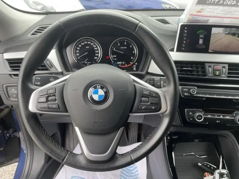 Vue intérieure centrée sur le volant BMW d’un X2 bleu métal 2021, avec tableau de bord et écran multimédia en arrière-plan.