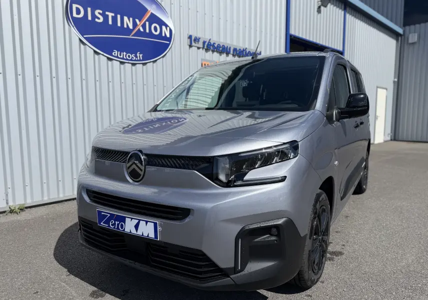 Vue 3/4 avant d'un Citroën Berlingo gris Artense métal avec calandre noire et jantes foncées, stationné devant un bâtiment.